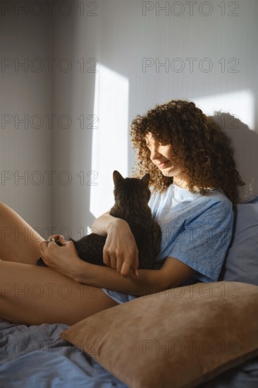 A woman with curly hair sits on a bed while holding a cat. Sunlight comes through the window, creating a warm atmosphere in the room. The moment shows a connection between the woman and her pet