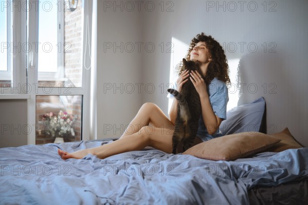 A woman in blue pajamas is sitting on a bed with blue sheets in a bright room. She is holding a cat and looking happy. Sunlight enters through the window and a small plant is seen outside
