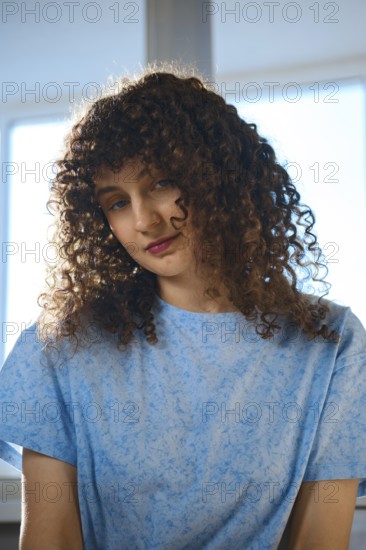 A woman with curly hair sits by a window. She wears a blue shirt and looks down with a thoughtful expression. The light from the window highlights her features and adds warmth to the scene