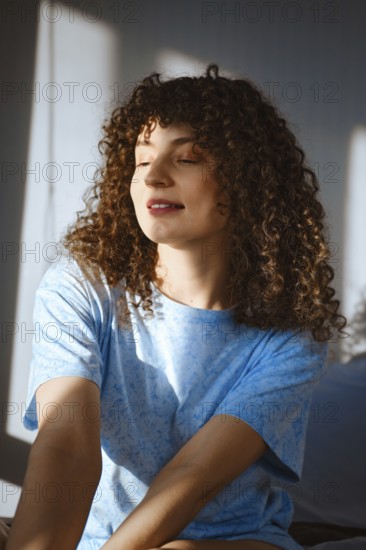 A woman with curly hair sits in a well-lit room. Sunlight streams through the window, creating warm shadows. She looks relaxed and happy, enjoying her moment in the soft light of the morning