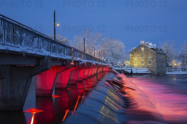Illuminated Moulin Neuf water flow control dam and walkway over Des Mille-Iles river plus New Mill on Ile des Moulins with first winter like snow in late autumn at dusk, Old Terrebonne, Quebec, Canada