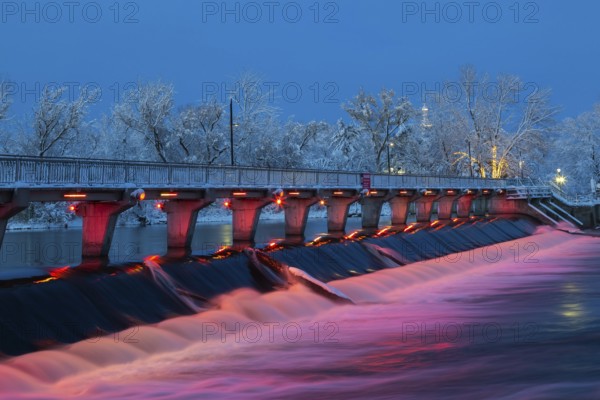 Illuminated Moulin Neuf water flow control dam and walkway over Des Mille-Iles river with first winter like snow in late autumn at dusk, Old Terrebonne, Quebec, Canada