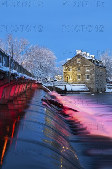 Illuminated Moulin Neuf water flow control dam and walkway over Des Mille-Iles river plus New Mill on Ile des Moulins with first winter like snow in late autumn at dusk, Old Terrebonne, Quebec, Canada
