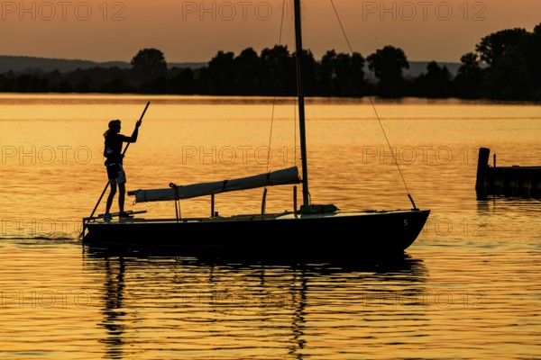 Sailing boat with person on calm Dümmer See brought into the harbor with a long stake at sunset, silhouette against orange sky, Lembruch, Dümmer See, Lower Saxony, Germany