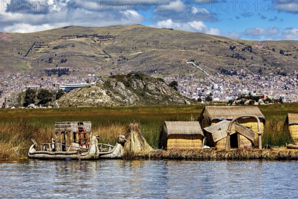 Reed houses and traditional lakeside boat under a cloudy sky with mountains, The floating reed islands of the Uros in Lake Titicaca in Peru