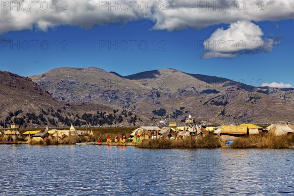 Scene with traditional huts on a lake against a mountain backdrop under blue skies and clouds, The floating reed islands of Uros in Lake Titicaca in Peru