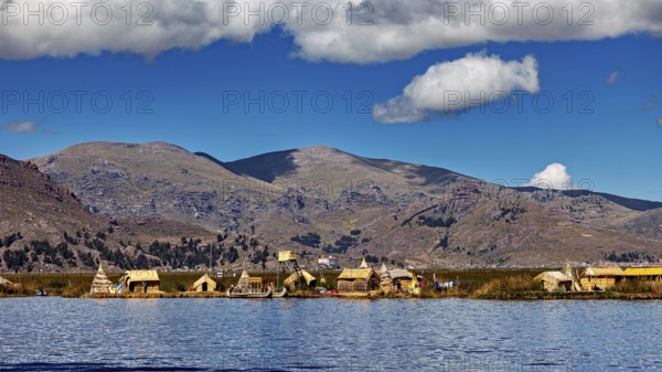 Row of reed huts on a calm lake under a partly cloudy sky and mountain backdrop, The floating reed islands of the Uros in Lake Titicaca in Peru
