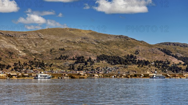 A calm lake in front of a mountainous landscape with boats and a village on the shore under a blue sky, The floating reed islands of the Uros in Lake Titicaca in Peru