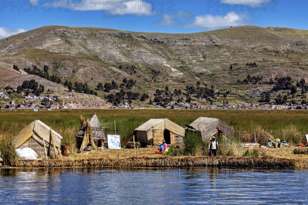 Huts on a reed island in front of a village and mountains, reflecting in the water, The floating reed islands of the Uros in Lake Titicaca in Peru