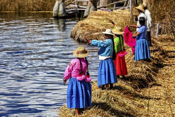 Women in colorful traditional clothing stand on the shore and look out at the water, the floating reed islands of the Uros in Lake Titicaca in Peru