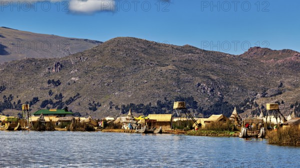 Huts on the shores of a lake in front of a mountainous landscape under a blue sky, The floating reed islands of the Uros in Lake Titicaca in Peru