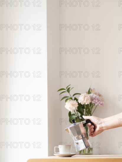 Woman is pouring coffee from moka pot into cup on wooden table with flowers in vase