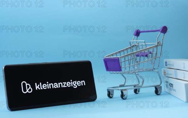 Dhaka, Bangladesh- 03 July 2025: Smartphone with Kleinanzeigen logo next to a small shopping cart and boxes against a blue background