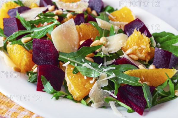 A salad made with chunks of beet, orange slices, and arugula. It is topped with cheese shavings and pine nuts, placed on a white plate with a striped napkin beside it