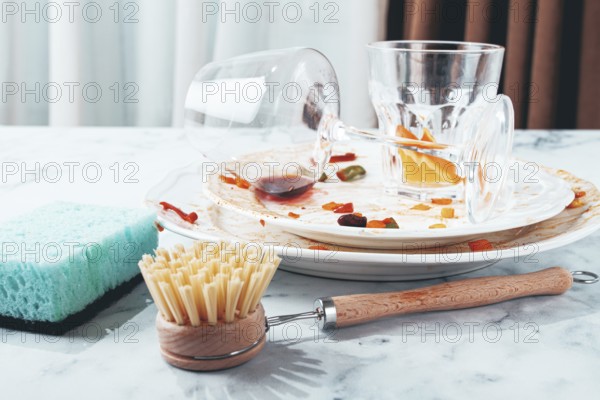 Dishes are stacked on a kitchen counter with food remnants after a meal. A cleaning brush and sponge are ready for washing the plates and glasses