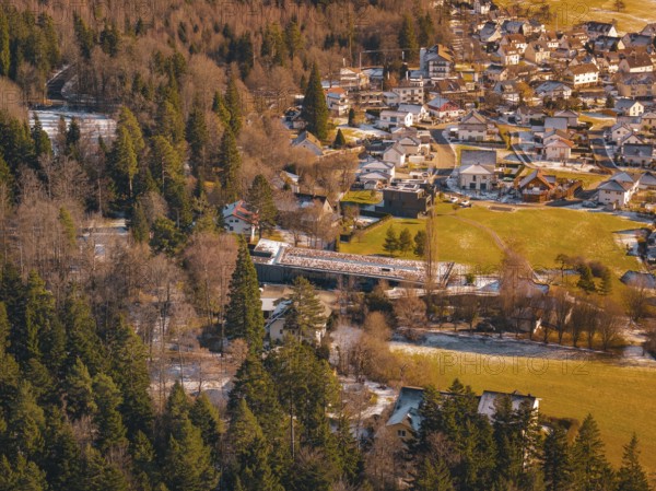 A bridge over a river surrounded by trees leads to a village in a snowy landscape, Hallwangen, Freudenstadt district, Germany