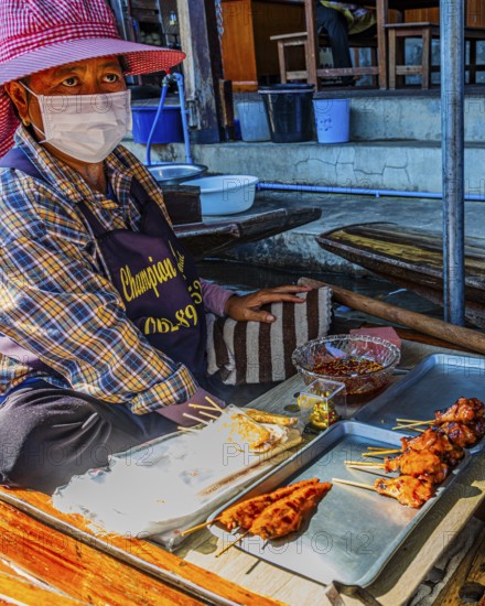 Elderly woman wearing mask sells barbecue skewers on her wooden boat on the Damneon Saduak floating markets channel, near Bangkok, Ratchaburi district, Thailand