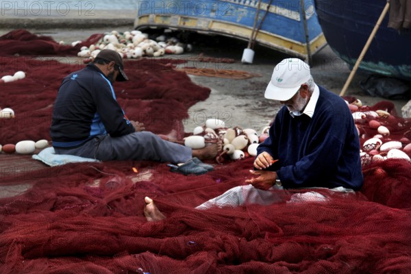 Fishermen repair nets in harbor surrounded by blue boats and tools, Essaouira, Marrakesh-Safi, Morocco
