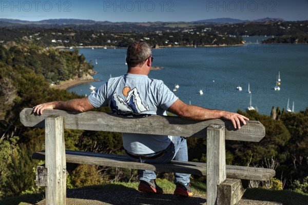 A man is sitting relaxed on a bench and enjoying the view of the Bay of Islands, Maiki, Bay of Islands, New Zealand