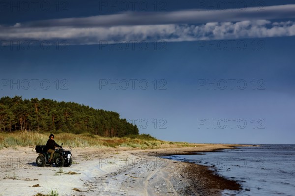 ATV rider rides along deserted coast with forest in background under clear sky, Kolka, Latvia