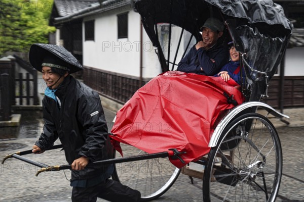 A rickshaw driver pulls two passengers through the streets of Kurashiki when it rains, Kurashiki, Japan