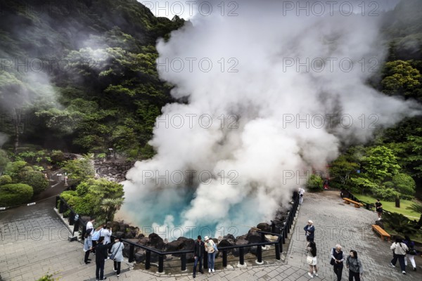 Group of visitors watching steaming blue lake surrounded by rocks and green foliage, Beppu, Oita, Japan