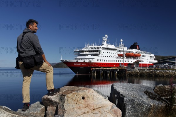 Man on rocks looking at a Hurtigruten ship in Kirkenes harbour, Kirkenes, Finnmark, Norway