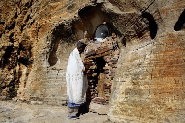 Priest stands in front of the rock church Abba Daniel Krokor in traditional clothing, zero