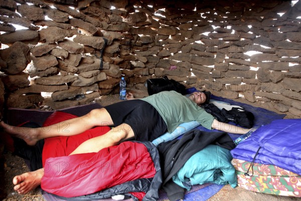 Person lying in a sleeping bag in a simple stone hut, zero