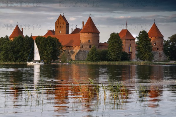 Trakai castle with red roof on a lake surrounded by trees and a sailboat, Trakai, Lithuania