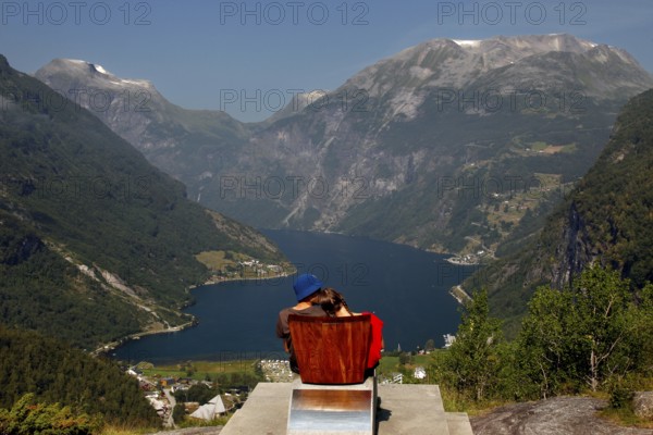 Tourist sits on a red chair and looks at the impressive panorama of Geirangerfjord, Flydalsjuvet, Geirangerfjord, Møre og Romsdal, Norway