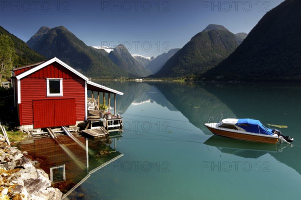 Red hut on the banks of Fjærlandsfjord near Mundal with boat and mountain backdrop, Mundal, Norway