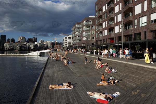 People relaxing on wooden walkway in the sun surrounded by new buildings, Oslo, Norway