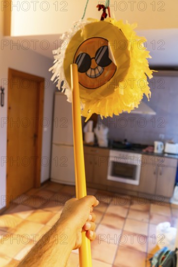 Hand holding yellow stick poised to strike a hanging emoji pinata in a home interior, playful birthday celebration scene with festive energy, candy and surprise imminent