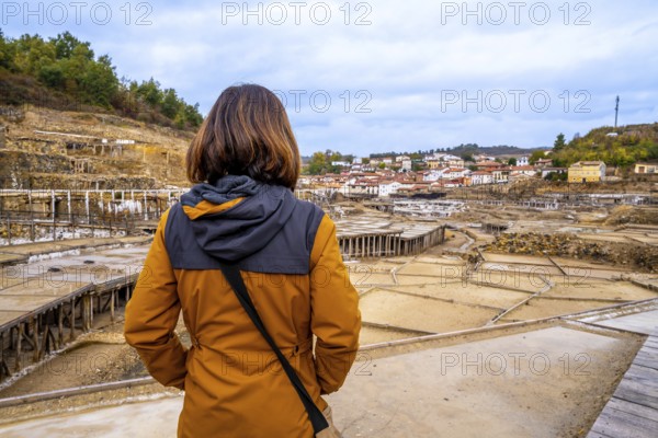 Woman tourist standing at anana salt valley, observing terraced evaporation ponds and traditional village houses against a rural basque landscape and cloudy autumn sky