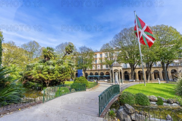 Gipuzkoa plaza park showing lush greenery, a pond with a small bridge, historic buildings, and the vibrant ikurrina flag flying proudly under a clear blue sky in san sebastian, basque country