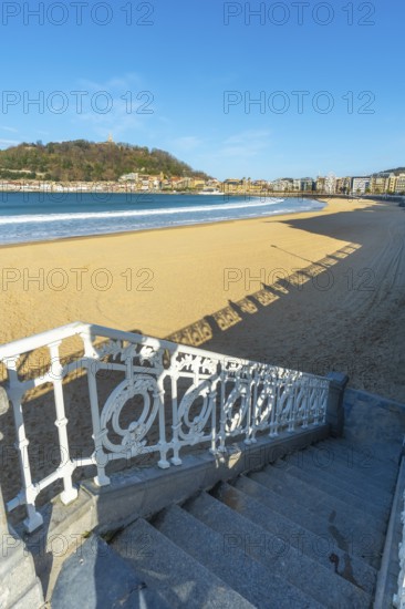 Scenic view looking down ornate white railings and stairs to the wide sandy beach of la concha bay, with the city and monte igueldo hill in the distance under a clear blue sky