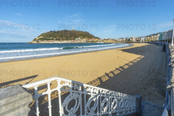 La concha beach view with its iconic promenade railing, golden sand and the city of san sebastian at the foot of mount urgull beneath a clear blue sky