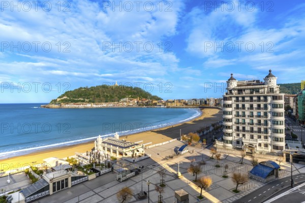 La concha beach bay stretching along the san sebastian cityscape and promenade, with urgull mountain and a hotel building under a blue sky with clouds