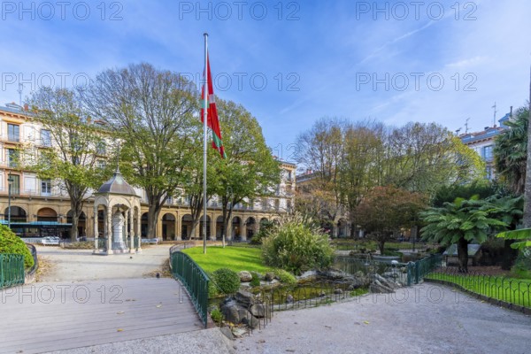 Gipuzkoa square in san sebastian showing the euskadi flag flying above the historic fountain and surrounding buildings with green trees under a clear blue sky