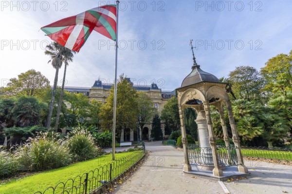 Basque ikurrina flag waving over historic gipuzkoa square in san sebastian, framed by lush gardens, pavilion, traditional buildings, pathways and sunny blue sky