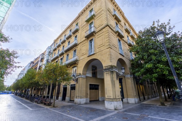Gipuzkoa square in san sebastian, basque country, with historic facades, wrought iron balconies and stone archways lining a tree shaded pedestrian street under blue sky