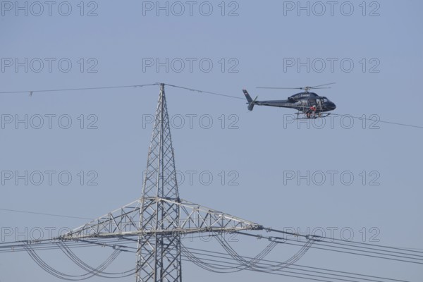 A helicopter carries out technical work on power lines next to a mast under a blue sky, an employee marks the top line of a new power line, Lemförde, Lower Saxony, Germany