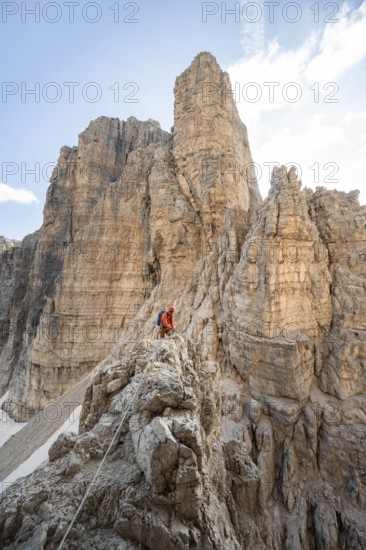 Mountaineer climbs an exposed rock in the secured Via Ferrata Bocciere Centrale via ferrata, spectacular rock towers in the back, Brenta Mountains, Brenta-Adamello Natural Park, Trentino, Italy