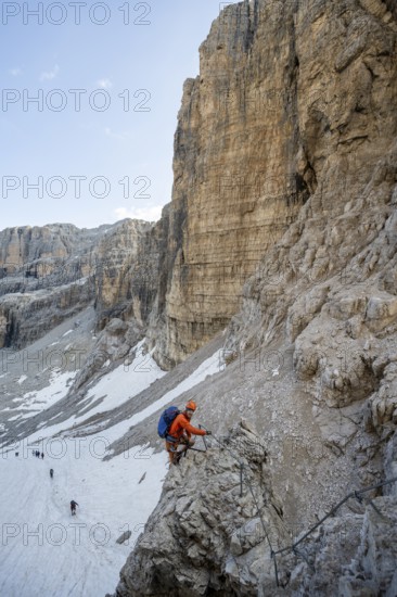 Mountaineer climbs on an exposed rock in the secured Via Ferrata Bocciere Centrale via ferrata, view of snow remains from the Vedretta del Sfulmini glacier, Brenta Mountains, Parco Naturale Brenta-Adamello, Trentino, Italy