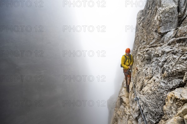 Mountaineers on a steep rock face on the Via Ferrata Oliva Detassis via ferrata in fog, steep mountains covered in clouds, Brenta Mountains, Trentino, Italy