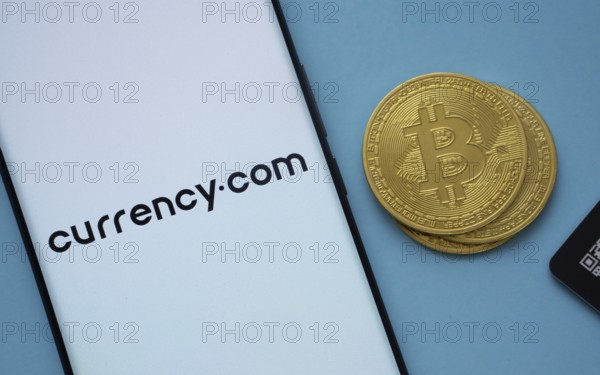 Dhaka, Bangladesh- 22 June 2025: Currency.com logo on smartphone screen near Bitcoin coin and QR code on blue background