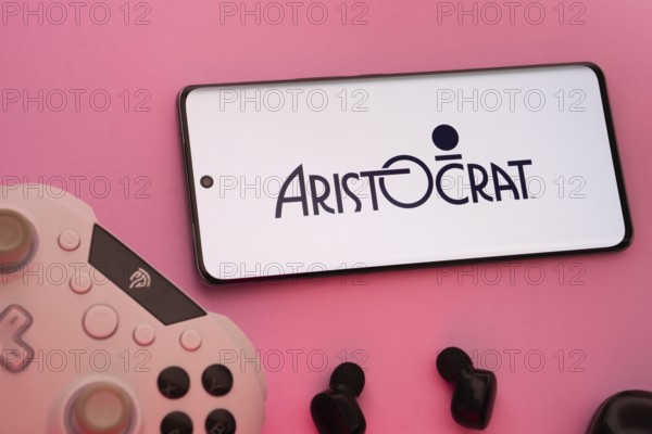 Dhaka, Bangladesh- 08 Apr 2025: A smartphone displaying the Aristocrat logo next to a gaming controller and earbuds on a pink surface