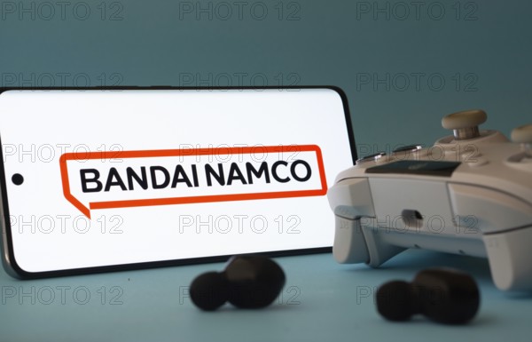 Dhaka, Bangladesh- 23 Mar 2025: A smartphone with a Bandai Namco Entertainment icon next to a controller and earbuds on a blue surface