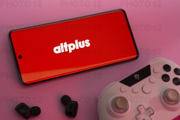 Dhaka, Bangladesh- 08 Apr 2025: A smartphone displaying the AltPlus Inc logo next to a gaming controller and earbuds on a pink surface